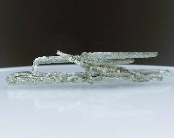 Può includere: Primo piano di un campione di minerale cristallino color argento. Il minerale ha un aspetto stratificato e strutturato con alcune piccole inclusioni marrone-arancio. Il campione è posto su una superficie bianca con uno sfondo scuro.