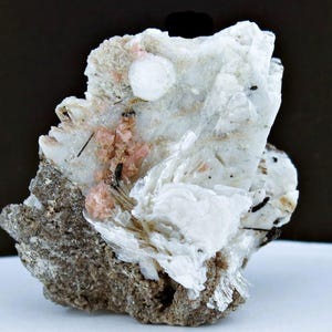 May include: A close-up of a mineral specimen featuring a mix of colors and textures. The rock has a rough, gray base with white and pink crystalline formations. Small black inclusions are scattered throughout the specimen.