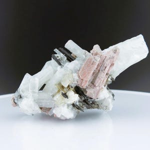 Könnte beinhalten: Ein Cluster aus weißen, rosa und schwarzen Kristallen. Die Kristalle sind in einem zufälligen Muster angeordnet und haben unterschiedliche Größen. Die Kristalle befinden sich auf einer weißen Oberfläche.