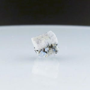 May include: A small, white, translucent crystal with blue and black inclusions. The crystal is on a white surface.