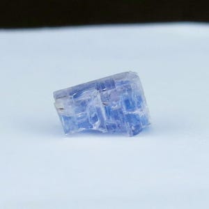 May include: A small, translucent, blue crystal cube. The crystal has a rough, textured surface and appears to be naturally formed. The cube is resting on a white surface, with a dark background.