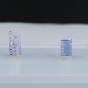May include: Two small, translucent blue-tinted crystal formations are displayed on a white surface against a black backdrop. The crystals have a rectangular shape with visible internal structures, suggesting a natural origin.