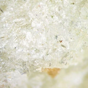 Op de afbeelding: Close-up van een getextureerd, gebroken wit oppervlak met kleine, donkere vlekjes en een paar kleine, kleurrijke deeltjes. Het oppervlak lijkt enigszins doorschijnend, met een subtiele, gelaagde uitstraling.