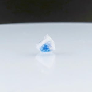 May include: A small, white crystal with a blue center. The crystal is sitting on a white surface.