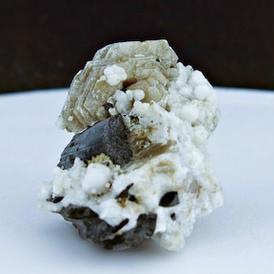 May include: A close-up of a natural mineral specimen. The rock features a combination of colors, including light brown, dark gray, and white. The texture is rough and crystalline, with small, rounded formations.