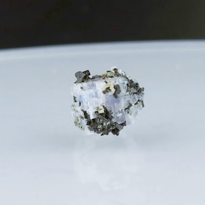 May include: A small, clear, blue crystal with black and white inclusions. The crystal is surrounded by a dark gray matrix.
