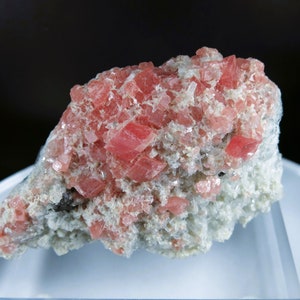 Puede incluir: Un cúmulo de cristales rosados y blancos sobre un soporte de exhibición blanco. Los cristales están dispuestos en un patrón aleatorio y tienen una textura rugosa y natural.