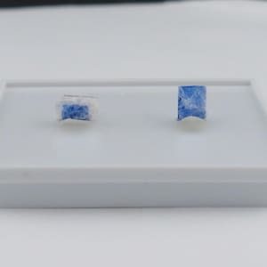 May include: Two small, rectangular stud earrings featuring a vibrant blue, crystalline stone set against a white, textured base. The earrings are displayed on a white plastic stand, showcasing their distinct blue color and natural, rough-cut texture.
