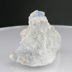 May include: A small, rough, white rock with a blue crystal on top. The crystal is about 1 cm in size and has a sharp, pointed shape.
