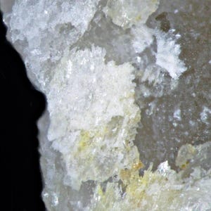 Könnte beinhalten: Nahaufnahme einer Mineralformation mit kristalliner Struktur. Das Bild zeigt eine Mischung aus weißen, durchscheinenden und gelblichen Farbtönen, mit einem dunklen schwarzen Hintergrund auf der linken Seite. Die Textur erscheint rau und uneben.