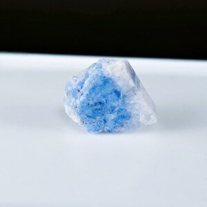May include: A blue and white crystal with a rough, textured surface. The crystal is a light blue color with white edges and is sitting on a white surface.