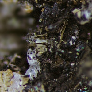 May include: Close-up of a mineral specimen with a rough, textured surface. The image shows a mix of dark, possibly metallic, and lighter, crystalline structures. Iridescent patches add color.