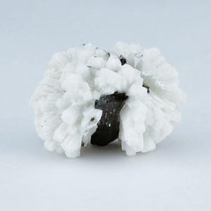 May include: A white mineral specimen with a spherical shape. The exterior is composed of numerous small, crystalline structures, creating a textured, almost fluffy appearance. A dark, possibly metallic, core is visible at the center.