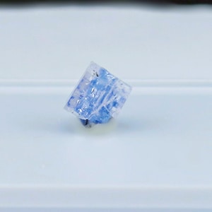 May include: A small, translucent, cubic crystal with a pale blue and white marbled appearance. The mineral specimen features sharp, geometric edges and a glossy, reflective surface texture, showcasing internal crystalline structures and variations.