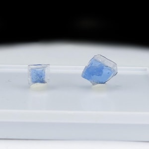 May include: Two small, translucent blue crystal fragments with irregular, angular shapes. The crystals feature a clear, glass-like outer layer surrounding a vibrant blue core, resting on a flat, white rectangular display base with raised edges.