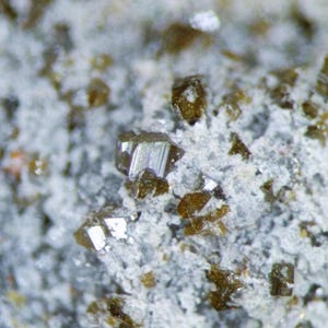 Puede incluir: Primer plano de una muestra mineral con pequeños cristales marrones incrustados en una matriz gris claro. Los cristales, con bordes afilados, tienen un aspecto metálico y reflectante.