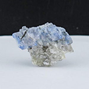 May include: A close-up of a mineral specimen with a layered appearance. The top layer is a vibrant blue, transitioning to a clear, crystalline base. The mineral sits on a white surface against a black backdrop.