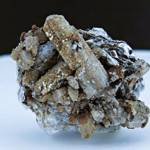May include: A close-up of a mineral specimen featuring a cluster of light brown, elongated crystals with white and brown speckling. The crystals are set against a backdrop of silver and grey mineral formations.