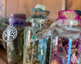 May include: Three clear glass bottles with cork stoppers. The left bottle has a silver tree charm and purple wax. The center bottle features green wax drips and a light stone. The right bottle is tied with twine and filled with dried rose petals.
