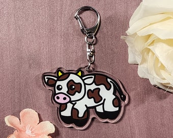 Könnte beinhalten: Ein klarer Acryl-Schlüsselanhänger mit einer Cartoon-Kuh mit braunen Flecken, schwarzen Hufen und gelben Hörnern. Die Kuh hat eine rosa Nase und ist an einem silberfarbenen Metallverschluss befestigt. Der Hintergrund ist mauvefarben.