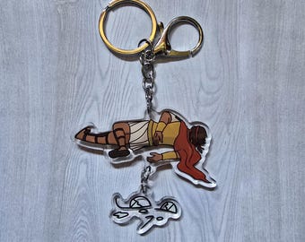 Könnte beinhalten: Ein klarer Acryl-Schlüsselanhänger mit einer Cartoon-Illustration einer Person in einem gelben und beigefarbenen Outfit mit roten Haaren. Ein kleinerer, passender Schildkrötenförmiger Anhänger hängt darunter. Der Schlüsselanhänger hat einen goldfarbenen Ring und Verschluss.