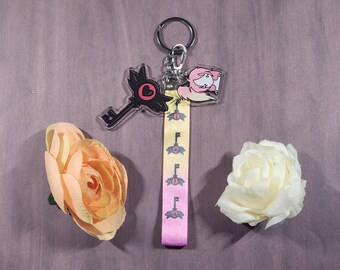 Könnte beinhalten: Ein rosa-schwarzer Schlüsselanhänger mit einem Herzdesign und einem Schlüssel, der an einem gelb-rosa Lanyard mit einem sich wiederholenden Schlüsselmuster befestigt ist. Das Lanyard ist an einem silbernen Schlüsselring befestigt.