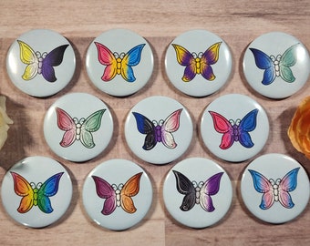 Könnte beinhalten: Zehn runde Anstecknadeln mit einem Durchmesser von 2,5 cm, die verschiedene farbige Schmetterlinge mit komplizierten Designs zeigen. Die Schmetterlinge repräsentieren verschiedene Pride-Flaggen.