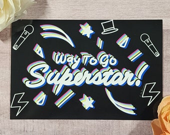 Könnte beinhalten: Schwarzer rechteckiger Aufkleber mit den Worten "Way to go Superstar!" in einem mehrfarbigen Design. Der Aufkleber zeigt weiße Strichzeichnungen eines Mikrofons, Sterne, einen Zylinder und Blitze.