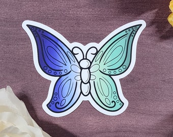 Könnte beinhalten: Ein blauer und grüner Farbverlauf-Schmetterlingsaufkleber mit weißem Umriss. Der Schmetterling hat ein detailliertes Design mit kleinen Kreisen an den Rändern der Flügel.