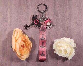 Könnte beinhalten: Ein rosa und schwarzer schlüsselförmiger Schlüsselanhänger mit einer Comicfigur darauf. Der Schlüsselanhänger hat ein rosa Band mit schwarzen Schlüsseln darauf gedruckt.