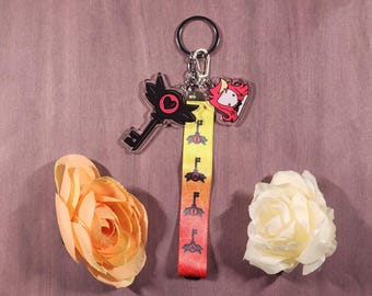 Könnte beinhalten: Ein schwarzer Schlüsselanhänger mit einem roten Herzen, einem rosa und roten Charakteranhänger und einem gelb-rot gestreiften Schlüsselband mit schwarzen Schlüsselmotiven. Die Anhänger sind an einem silbernen Schlüsselring befestigt.