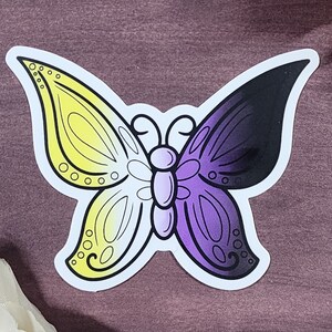 May include: A sticker of a butterfly with yellow, white, purple, and black wings. The butterfly is a symbol of the non-binary gender identity.