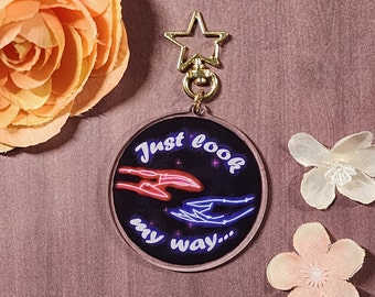 Könnte beinhalten: Ein runder, transparenter Acryl-Schlüsselanhänger mit schwarzem Hintergrund und neonrosa und blauen Händen, die sich gegenseitig zuwenden. Der Text "Just look my way..." ist in Weiß auf dem Schlüsselanhänger geschrieben.