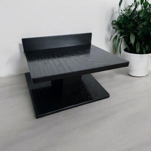 May include: A black wooden display stand featuring a square base, a central support pillar, and a square top platform with a small vertical back ledge. The wood grain is visible through the dark finish, creating a textured appearance.