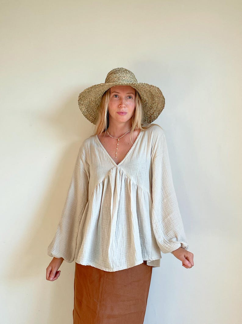 May include: A woman wearing a cream-colored, loose-fitting, long-sleeved blouse with a V-neck and a brown skirt. She is wearing a wide-brimmed straw hat.