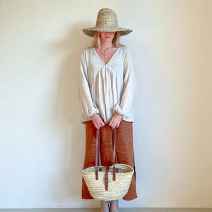 May include: A woman wearing a white linen blouse, a brown skirt, and a straw hat is holding a woven basket bag with leather straps. She is wearing blue espadrille sandals.