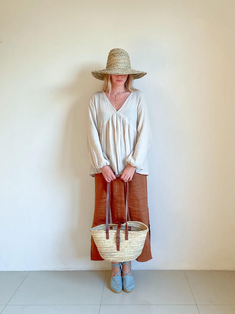 May include: A woman wearing a white linen blouse, a brown skirt, and a straw hat is holding a woven basket bag with leather straps. She is wearing blue espadrille sandals.