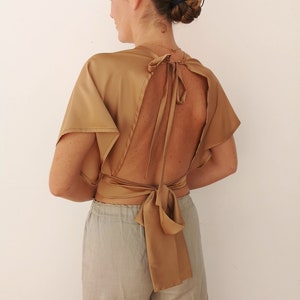 Pu&ograve; includere: Un top in satin dorato con schiena scoperta e un nodo sul retro. Il top &egrave; indossato con pantaloni grigi chiari.