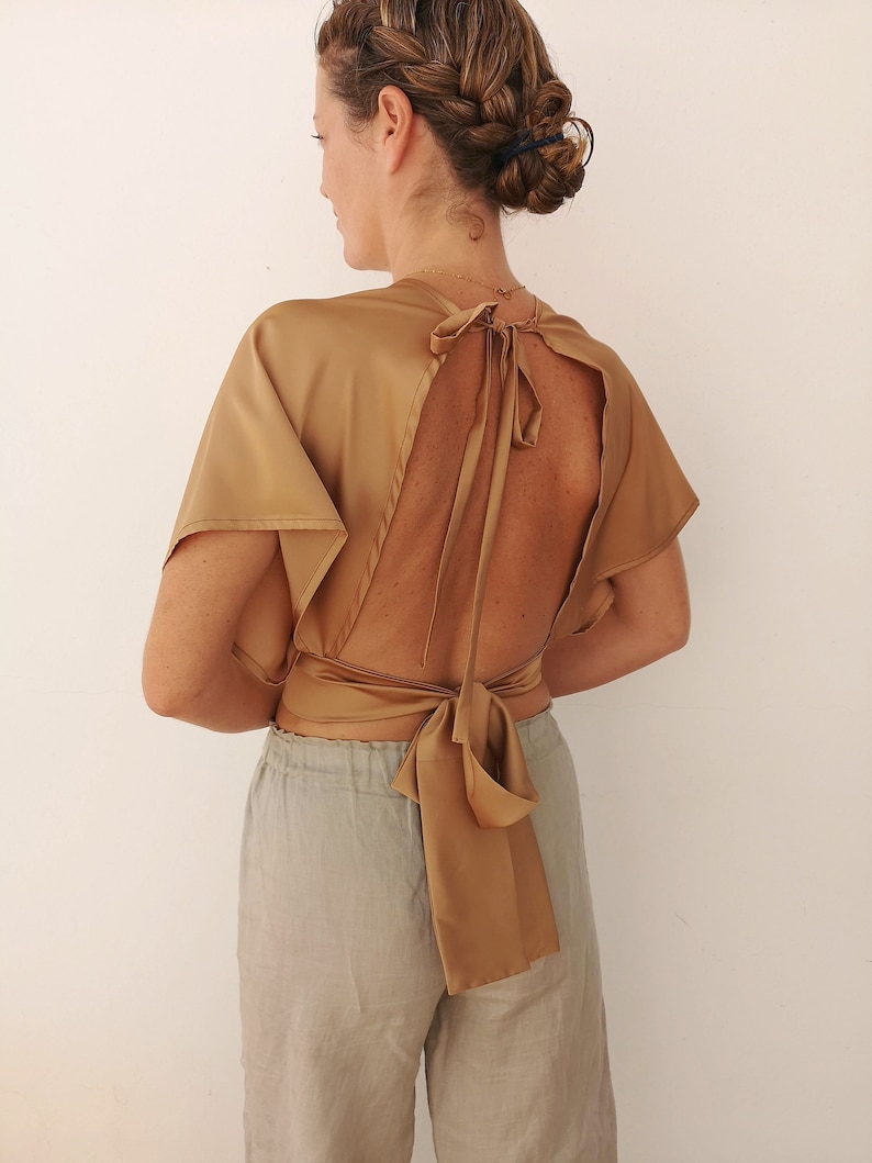 Pu&ograve; includere: Un top in satin dorato con schiena scoperta e un nodo sul retro. Il top &egrave; indossato con pantaloni grigi chiari.