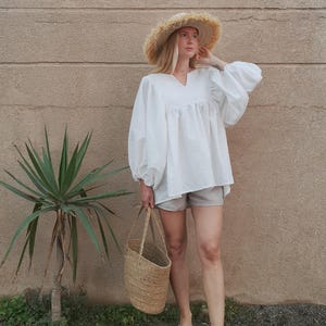 May include: A woman wearing a white, loose-fitting blouse with a V-neckline and long sleeves. She is also wearing light gray shorts, a straw hat, and sandals. She is carrying a woven tote bag.