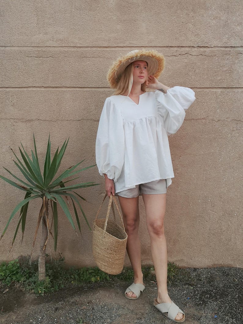May include: A woman wearing a white, loose-fitting blouse with a V-neckline and long sleeves. She is also wearing light gray shorts, a straw hat, and sandals. She is carrying a woven tote bag.