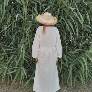 May include: A white maxi dress with long sleeves and a gathered waist. The dress is worn by a person wearing a straw hat and brown boots. The person is standing in front of a green leafy background.