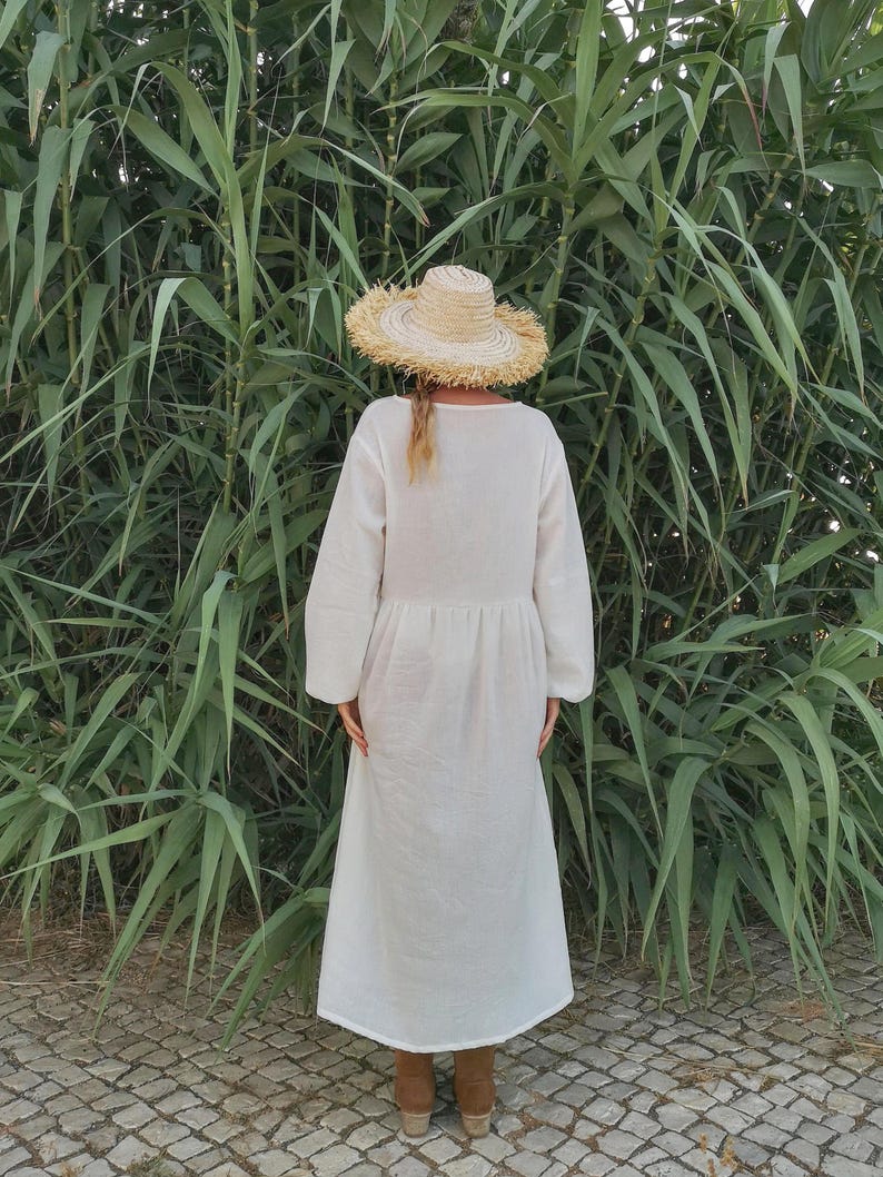 May include: A white maxi dress with long sleeves and a gathered waist. The dress is worn by a person wearing a straw hat and brown boots. The person is standing in front of a green leafy background.
