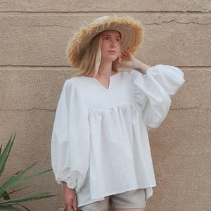 May include: A white, loose-fitting blouse with long, puffy sleeves and a V-neckline. The blouse is worn with light gray shorts and a straw hat.
