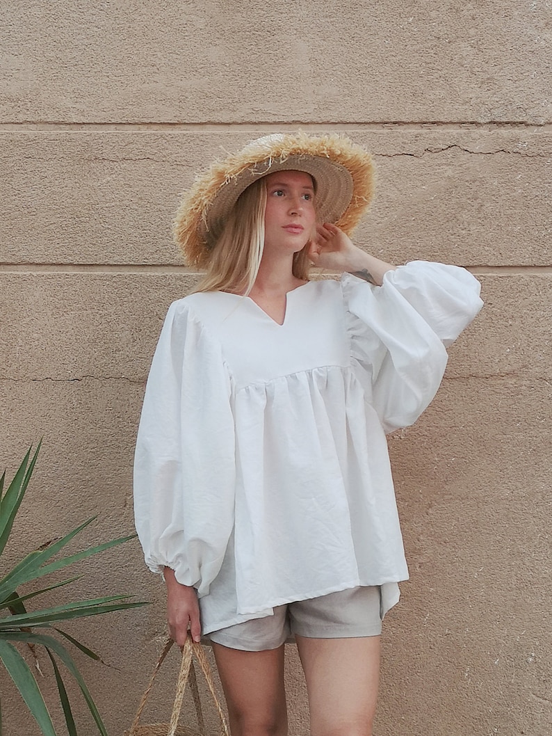 May include: A white, loose-fitting blouse with long, puffy sleeves and a V-neckline. The blouse is worn with light gray shorts and a straw hat.