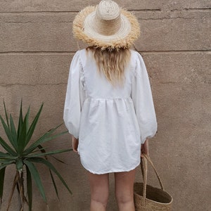 May include: A white linen shirt with a gathered waist and long sleeves. The shirt is worn with a large, straw hat with a fringe trim and a straw tote bag.