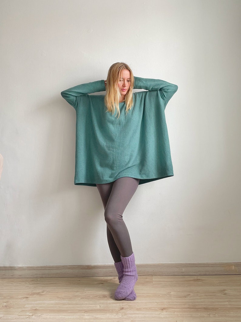 May include: A woman wearing a teal oversized sweater with long sleeves and grey leggings. She is wearing purple socks and has her hands on her head.