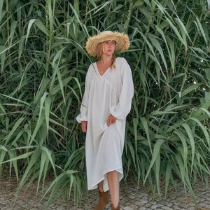 May include: A woman wearing a white maxi dress with a wide, floppy straw hat. She is standing in front of a wall of green plants.