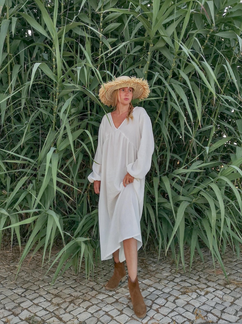 May include: A woman wearing a white maxi dress with a wide, floppy straw hat. She is standing in front of a wall of green plants.