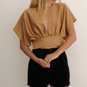 Pu&ograve; includere: Una donna indossa una camicetta di raso marrone con una profonda scollatura a V e maniche corte. Indossa anche pantaloncini neri.
